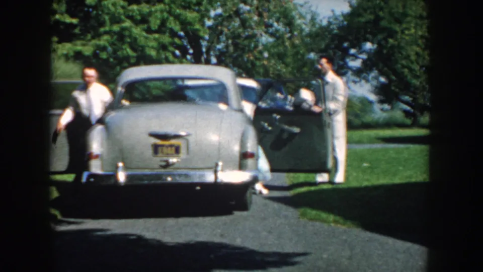 Two dressed men stand beside the stopped gray sedan in a 1959 Nebraska home-movie view of a garden arrival.