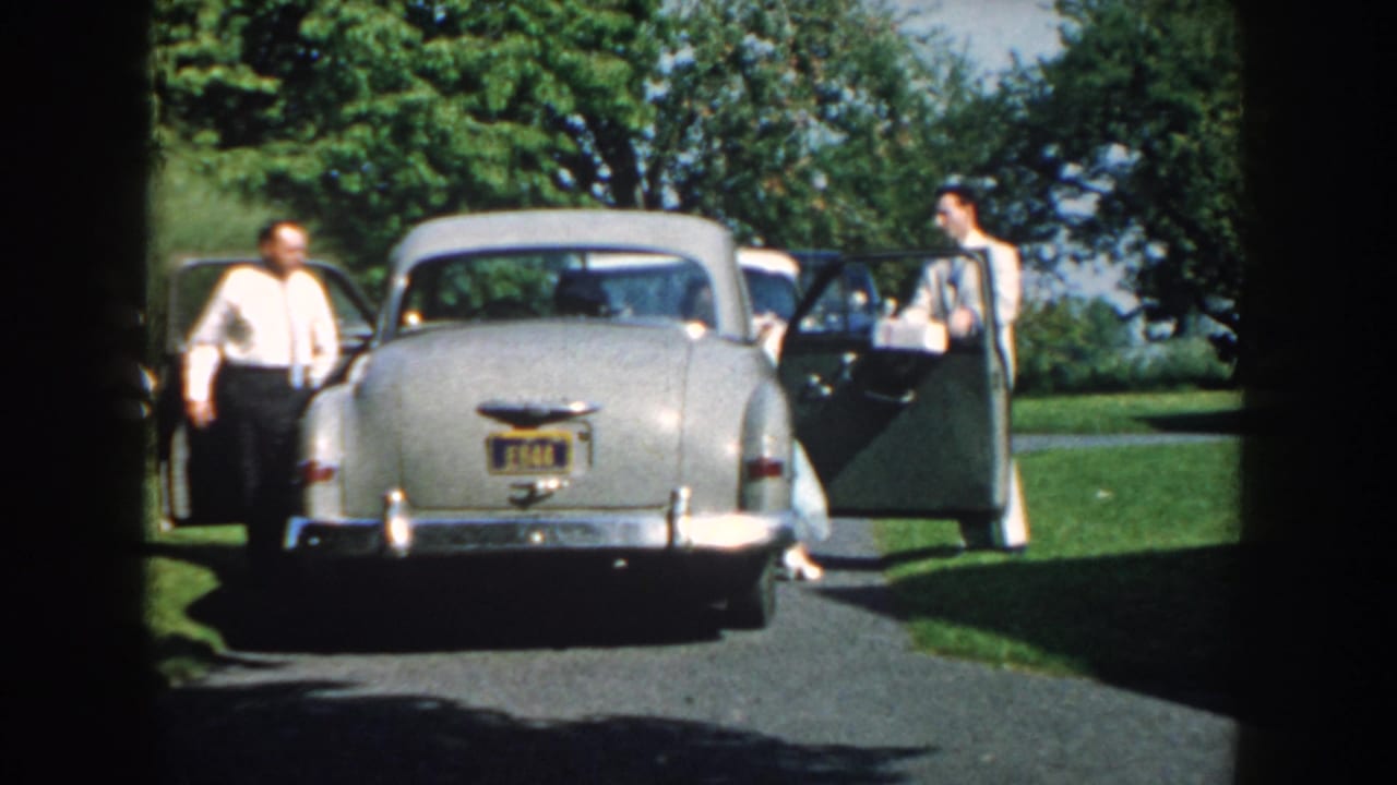 Two men stand near a silver sedan with both side doors open on a paved drive.