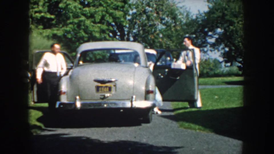 In 1959 Nebraska home-movie footage, two formally dressed men stand by an open silver sedan in a green outdoor setting.