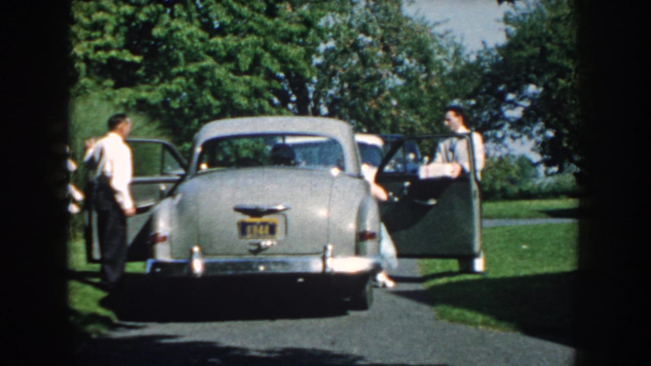 The silver 1950s sedan remains parked as two men in formal clothing stand near opposite open doors outdoors in Nebraska.