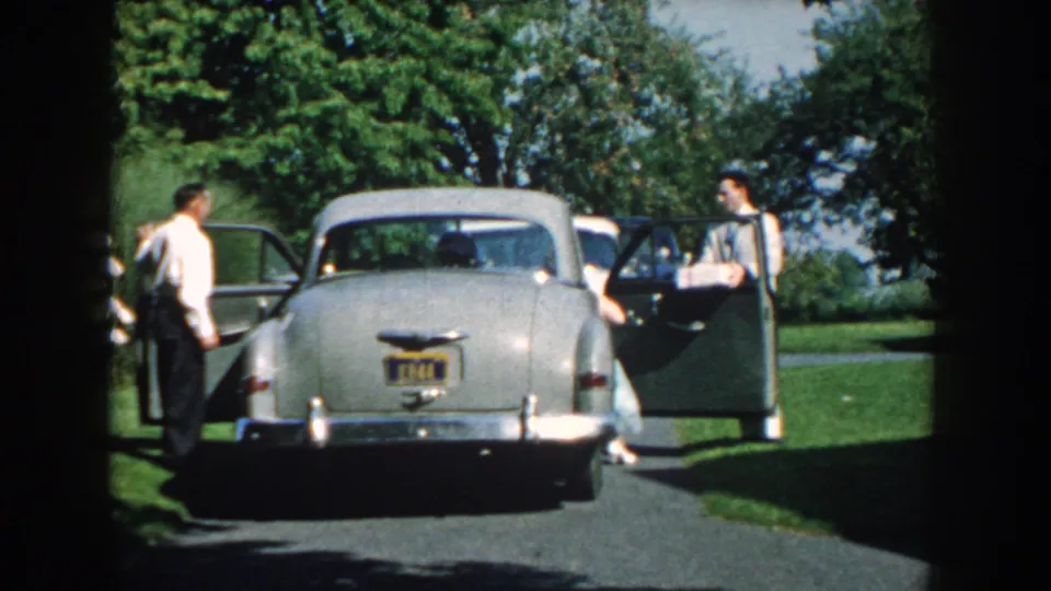 The silver 1950s sedan remains parked as two men in formal clothing stand near opposite open doors outdoors in Nebraska.