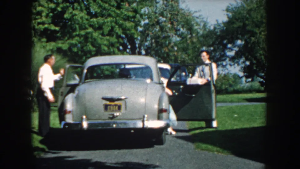 A formally dressed man gestures or steps near the sedan as another man stands by the open passenger-side door in the outdoor scene.