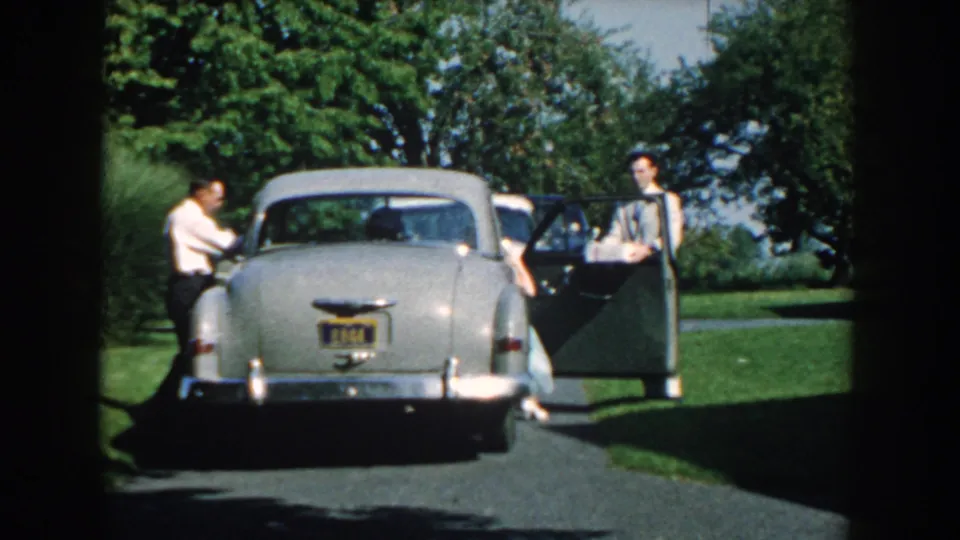 The 1959 Nebraska home-movie frame shows two formally dressed men beside a parked silver sedan with open doors and trees behind them.
