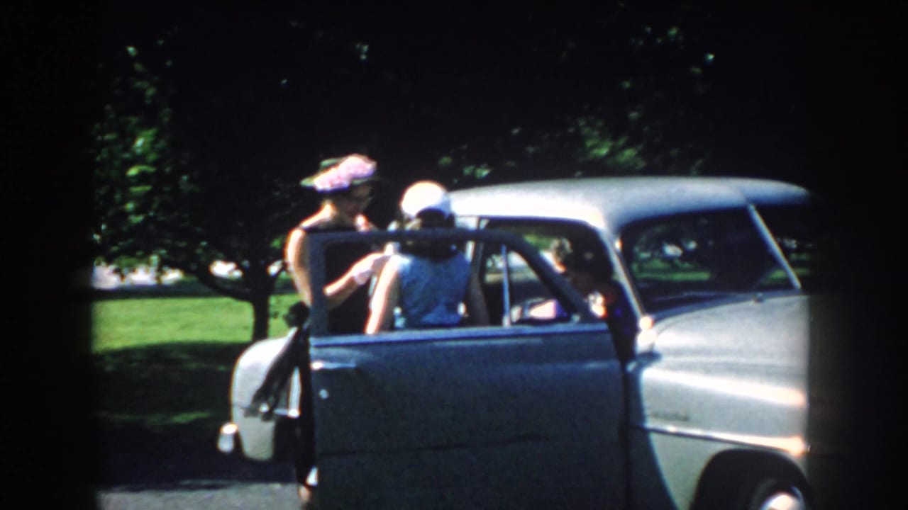A woman stands outside a vintage car while another is at the open door.