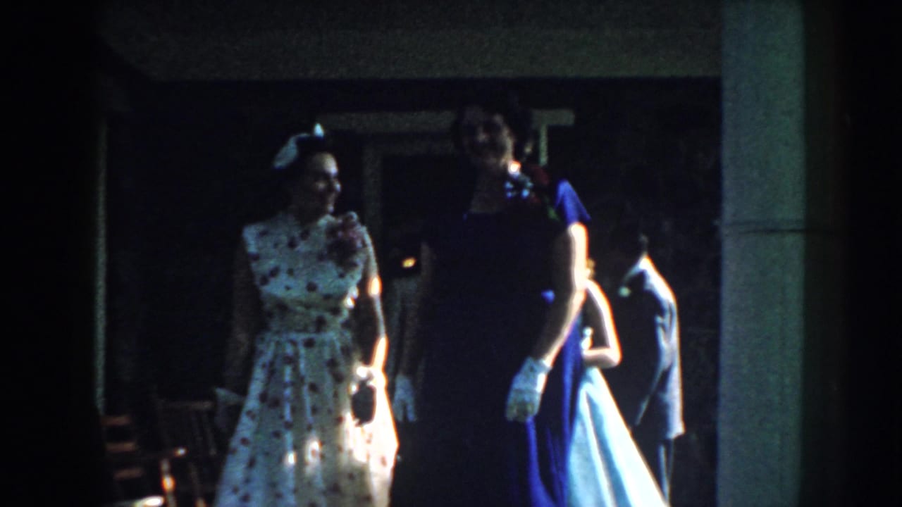 Two well-dressed women walk outside near a doorway and porch column.