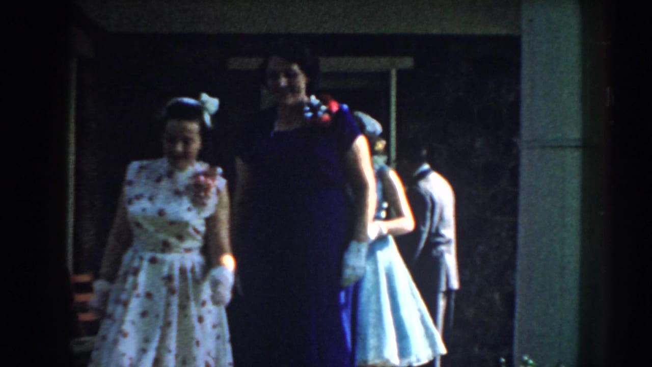 Women in formal dresses walk past a house entrance with other guests behind them.