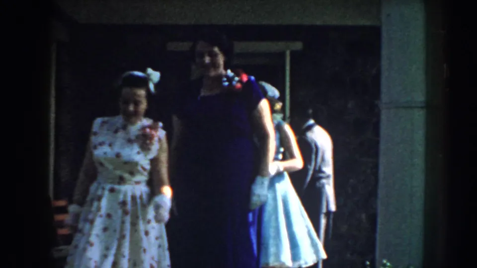 Formally dressed women walk outside a Nebraska house in 1959, with gloves, patterned fabric, and other suited guests partly visible behind them.