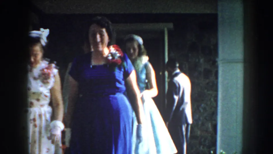 A woman in a blue dress and white gloves moves close to the camera, with another woman and suited figures near the porch in 1959 Nebraska footage.