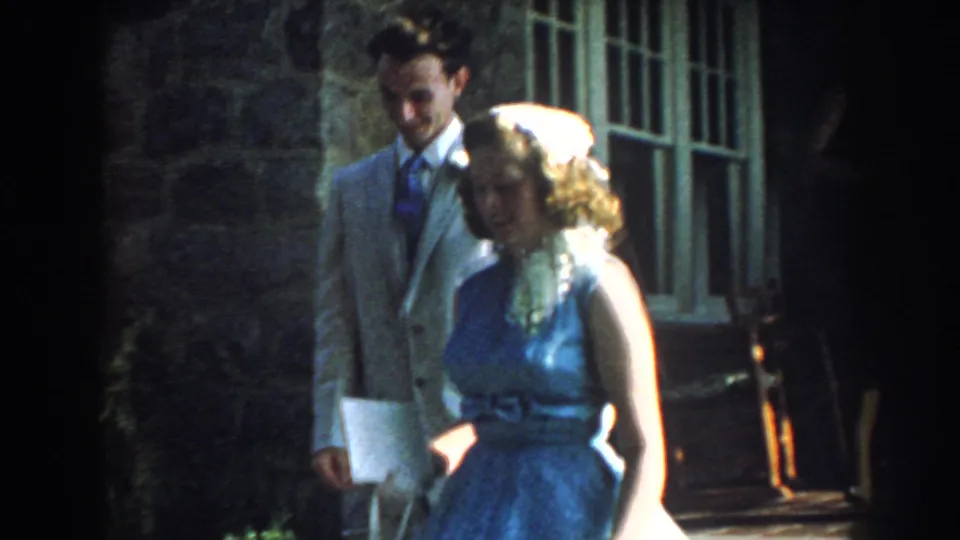 A man in a light suit and tie stands near a woman in a blue dress by a stone wall and window in 1959 Nebraska home-movie footage.