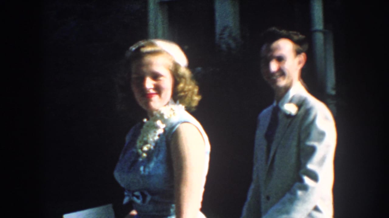 Smiling woman in a light dress and man in a light suit outside a shaded stone house.