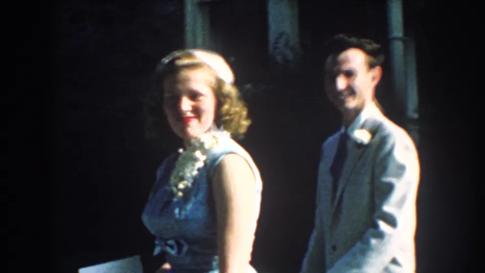 A smiling woman with a white headpiece and floral accessory stands near a man in a light suit outside the Nebraska stone house.