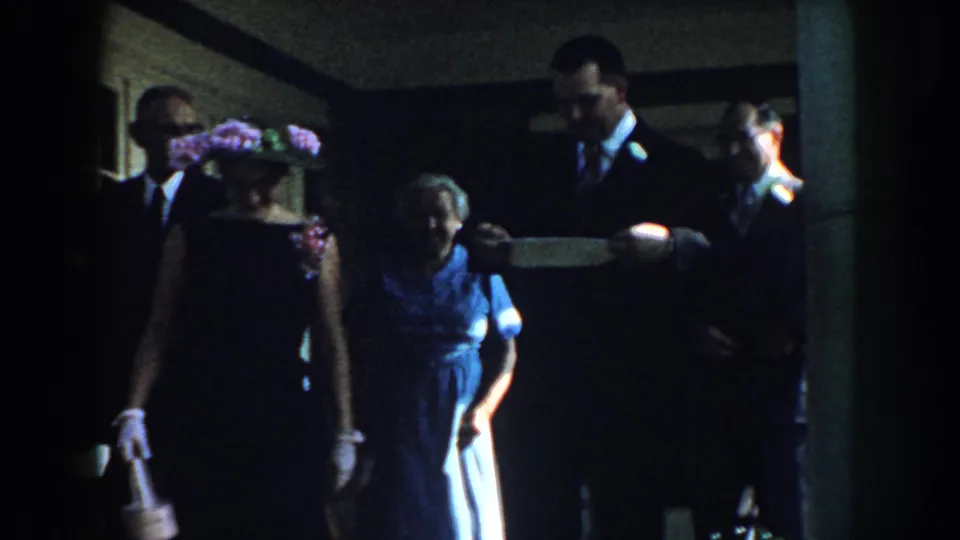 Several adults in 1959 Nebraska formalwear stand on a porch, including a woman in a floral hat with a small basket.