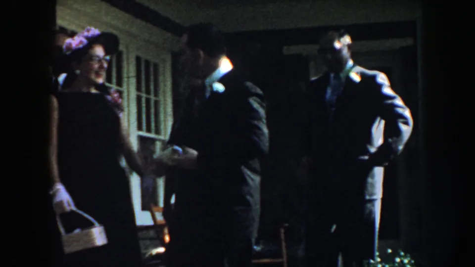 Adults in suits and dresses stand on a Nebraska porch, with a floral headpiece, basket, boutonnieres, and house windows visible.