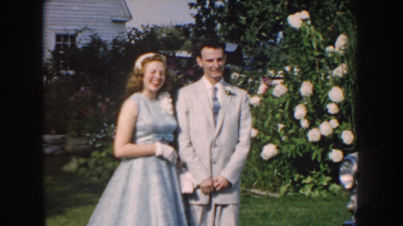 In 1959 Nebraska home-movie footage, a young couple poses outdoors in wedding attire near a lawn, white roses, and a white house.