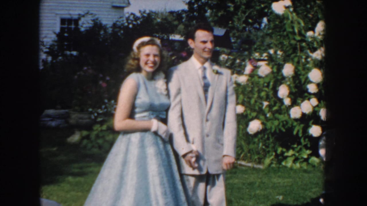 Smiling couple poses on a lawn with white roses and a house in the background.