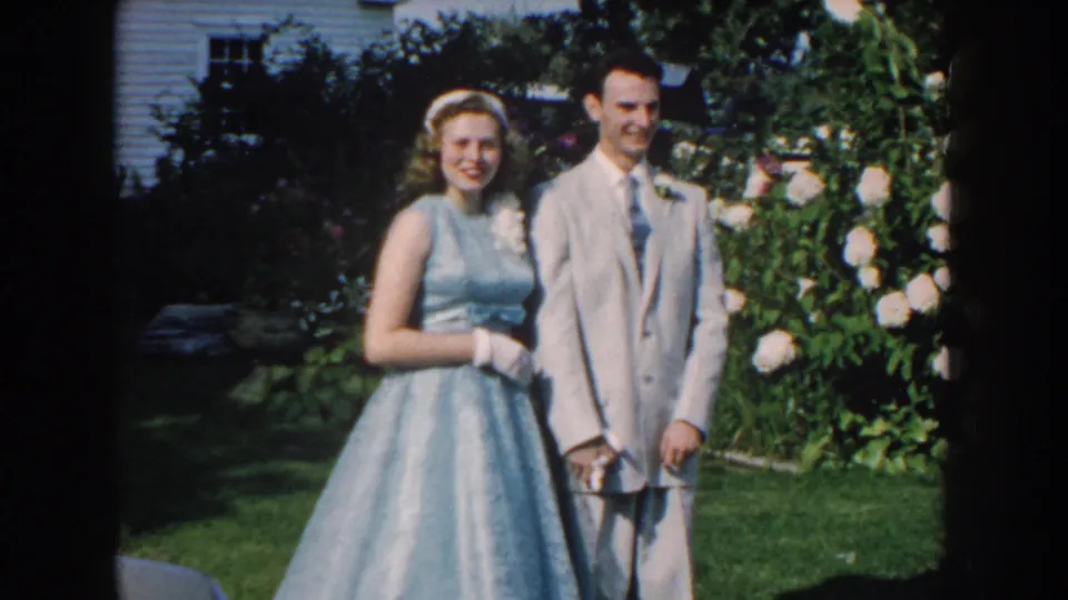 A 1959 Nebraska wedding couple poses in bright outdoor light, with white roses and a house partly visible behind them.
