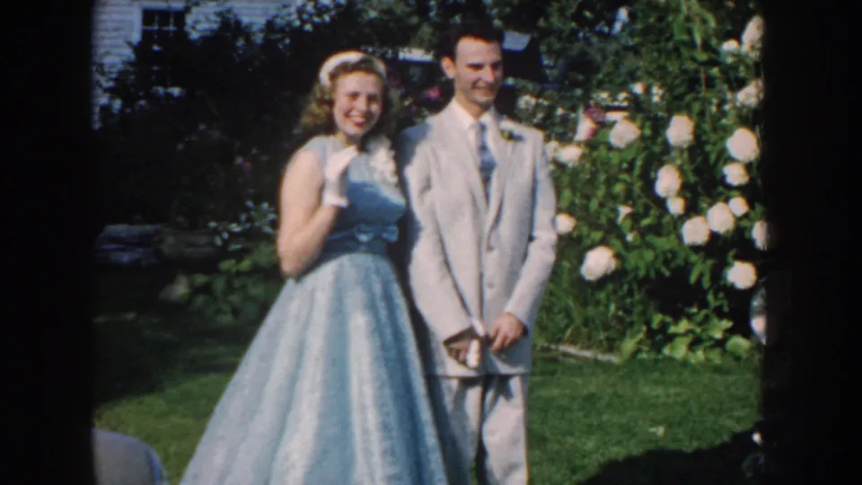 The woman lifts a gloved hand in a small wave as the man stands beside her in the 1959 Nebraska garden wedding scene.