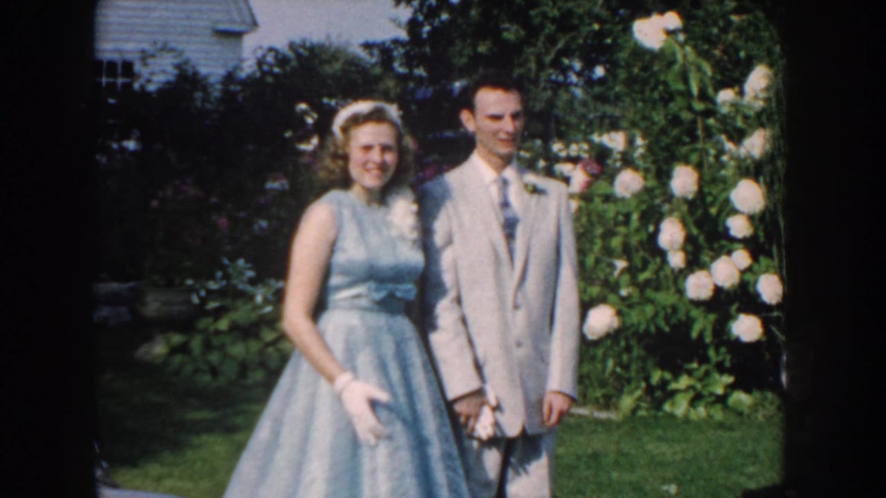 In 1959 Nebraska home-movie footage, a young woman in a blue dress and a young man in a light suit pose outdoors by white blossoms.
