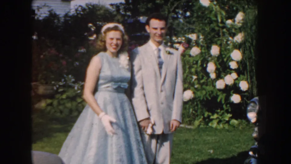 A soft 8mm frame shows the formally dressed couple facing the camera outdoors, with garden greenery and white blossoms behind them.