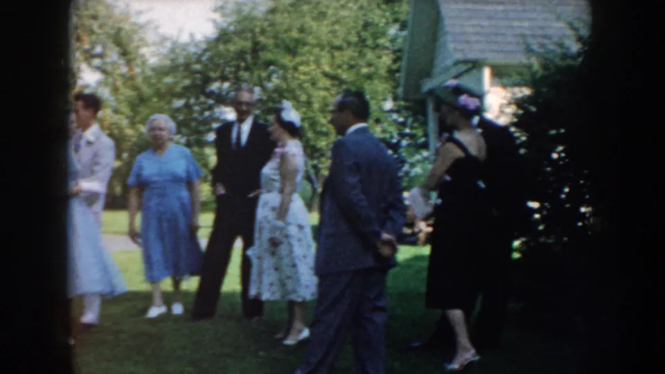 A wider 1959 Nebraska home-movie view shows adults gathered outdoors on a lawn, with some in suits and dresses near garden plantings.