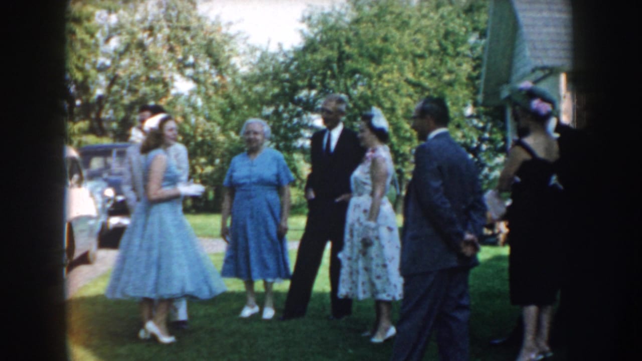 Nebraska 1959 home-movie frame showing dressed wedding guests gathered on a lawn beside mid-century cars.