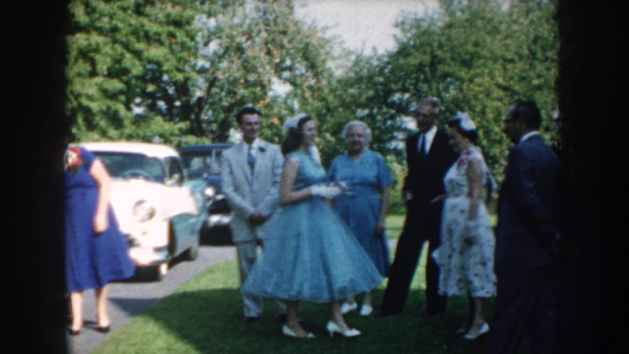 Group of wedding guests stands on grass beside a driveway with cars behind them.