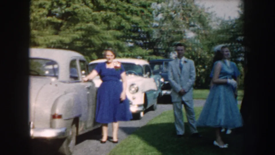 Nebraska 1959 wedding scene with a woman in a blue dress beside a gray car and guests in formalwear nearby.