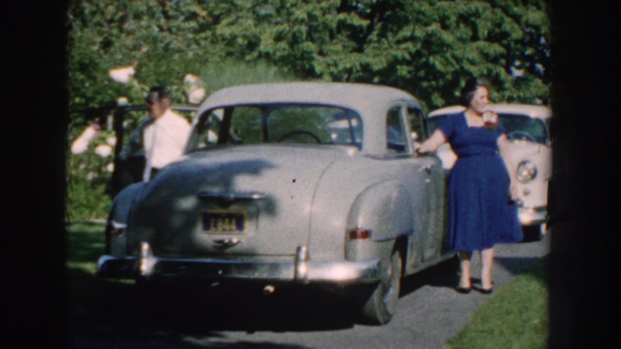 Woman in blue dress stands beside a silver sedan on a sunny driveway.