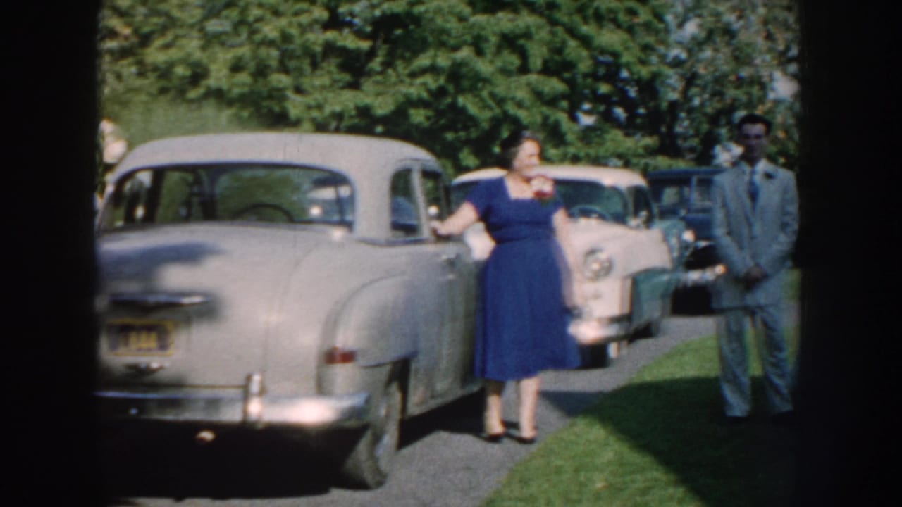 1959 Nebraska outdoor gathering with a woman in a blue dress near classic cars and a man in a light suit on the lawn.