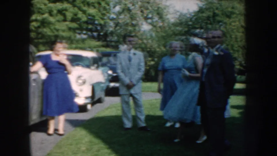 1959 Nebraska home-movie frame of adults in dresses and suits congregating outdoors beside parked mid-century cars.
