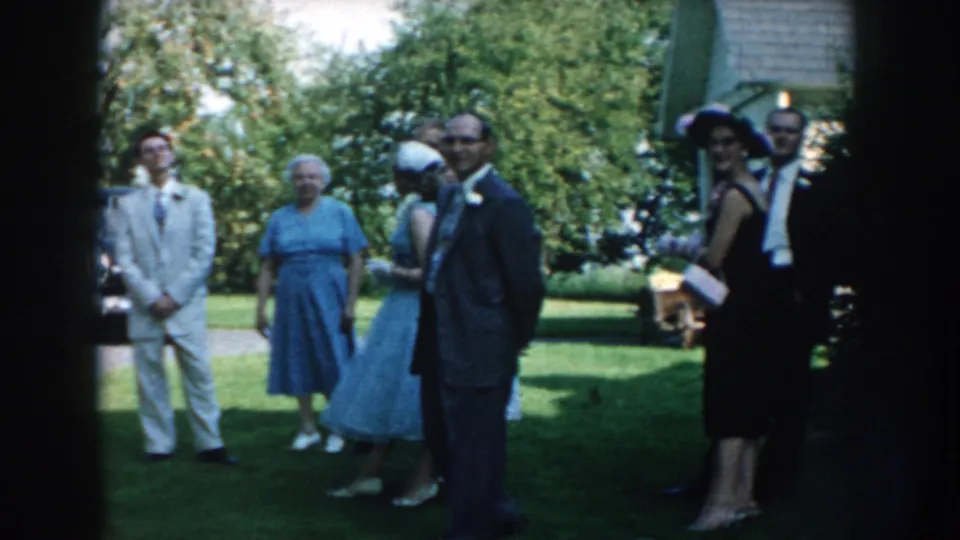 1959 Nebraska outdoor gathering with adults in formal attire standing on a green lawn near trees and a building edge.