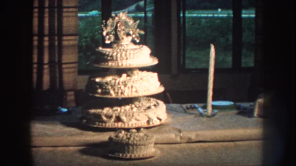 Final frame centers the ornate white wedding cake on its table, with curtains and windows behind it in the home-movie scene.