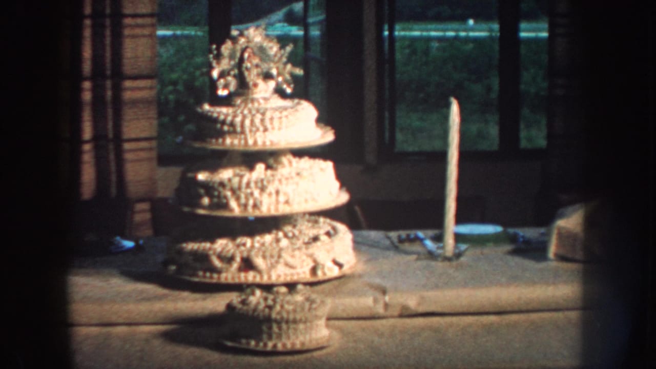 1959 Nebraska 8mm frame showing a multi-tiered white wedding cake on a table with a tall candle and curtained window behind it.