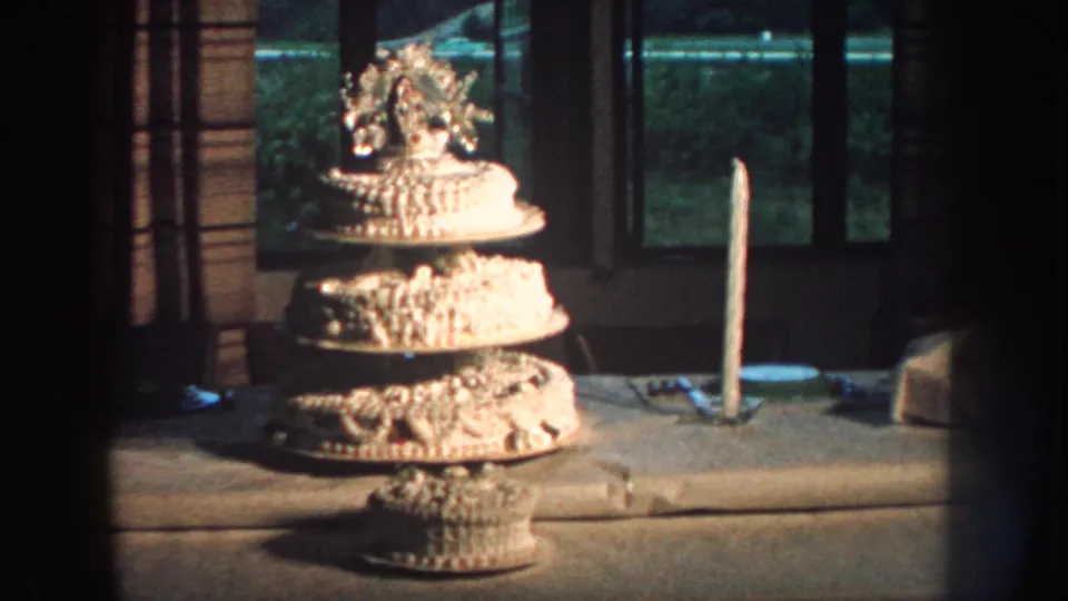 1959 Nebraska 8mm frame showing a multi-tiered white wedding cake on a table with a tall candle and curtained window behind it.