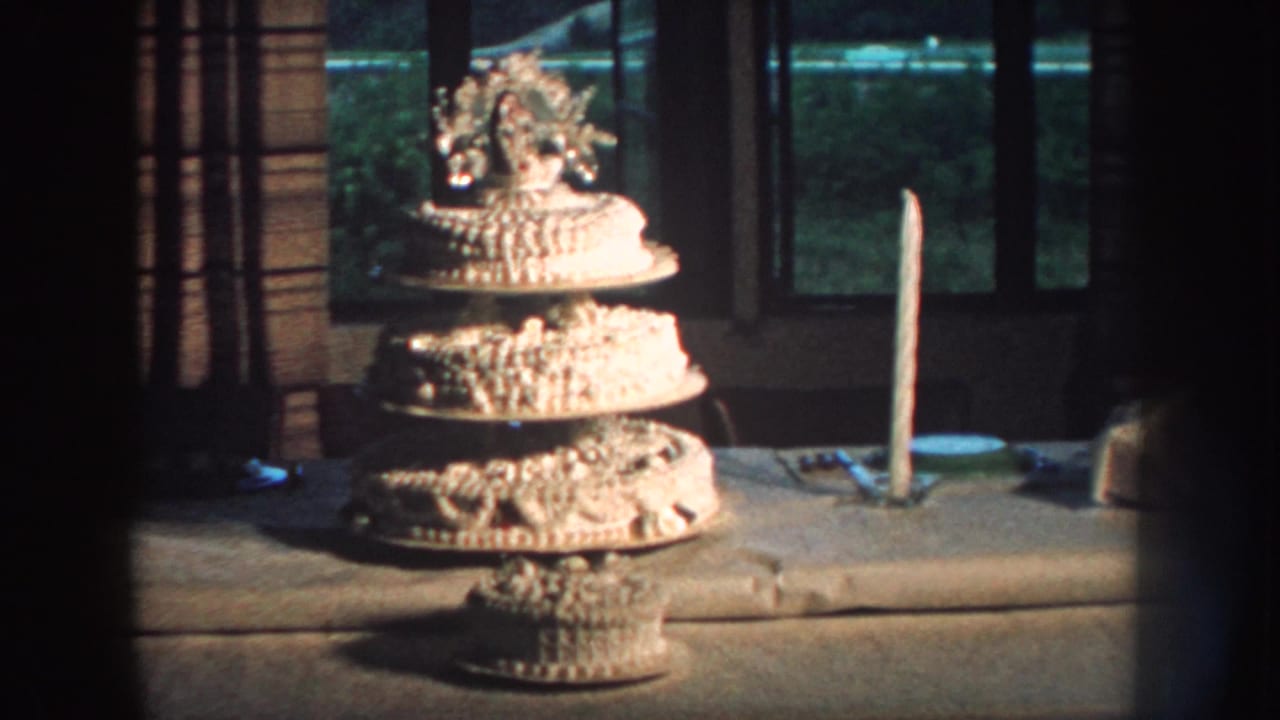 1959 Nebraska home-movie frame of a decorated wedding cake on a tiered stand, with a single candle and dark window backdrop.