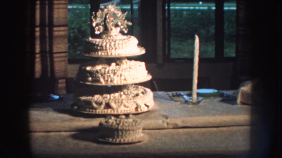 1959 Nebraska 8mm frame showing the wedding cake from a similar angle, with soft grain, a candle, curtains, and greenery outside.