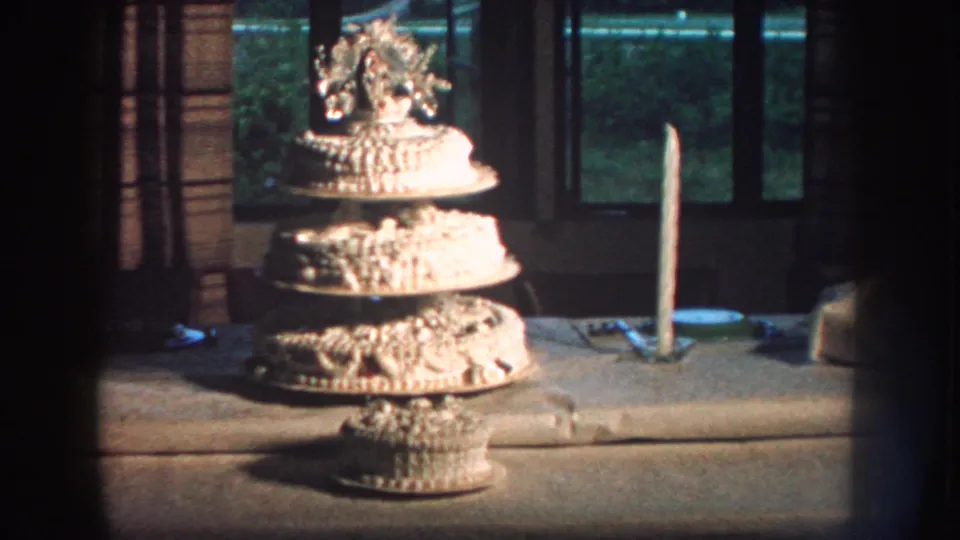 1959 Nebraska home-movie frame of a tiered wedding cake on a table, with a candle beside it and window panes behind.