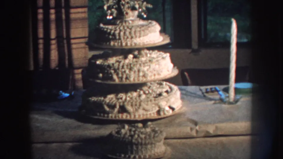 1959 Nebraska 8mm frame with a white tiered wedding cake, ornate frosting, a topper, and a tall candle near curtained windows.