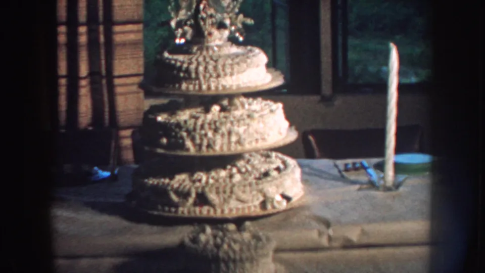 1959 Nebraska 8mm frame showing a decorated tiered wedding cake on a table, with a tall white candle and curtained window in the background.