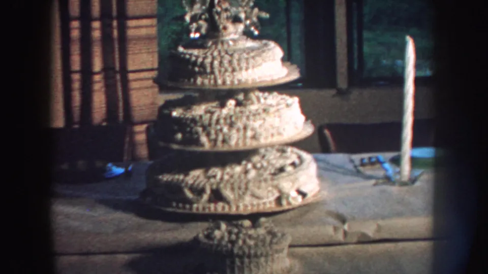 1959 Nebraska 8mm frame of a decorated white wedding cake with topper, placed indoors beside a tall candle and curtained window.