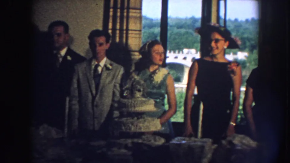 Nebraska 1959 home-movie frame showing wedding guests behind a white tiered cake in an indoor reception room.