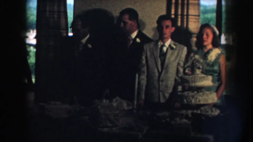 Nebraska 1959 home-movie still showing reception guests behind a white cake, partly obscured by a foreground silhouette.