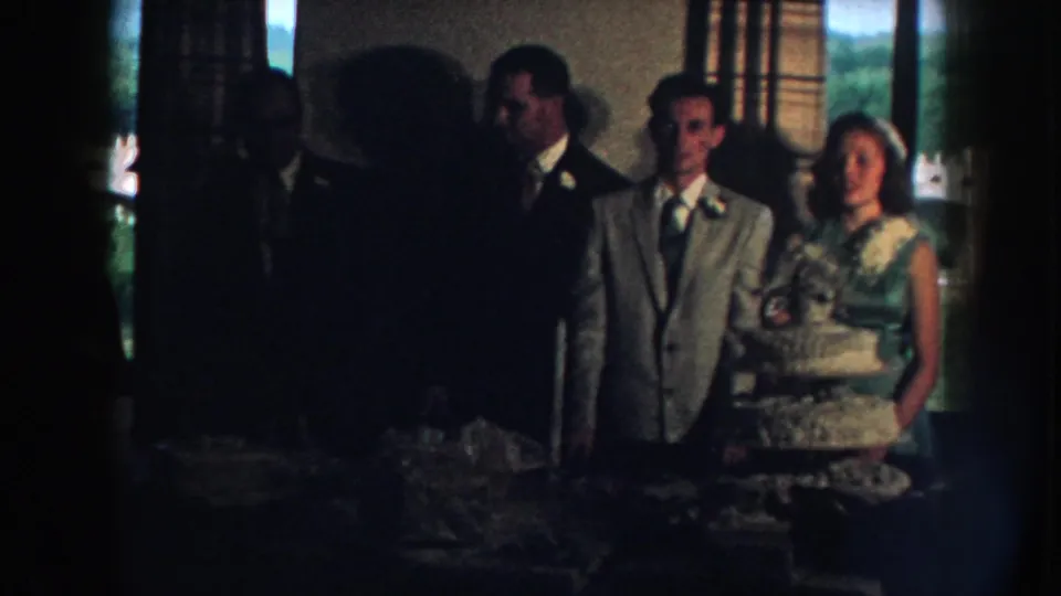 1959 Nebraska home-movie frame showing a wedding party posed near a tiered cake inside a private reception room.