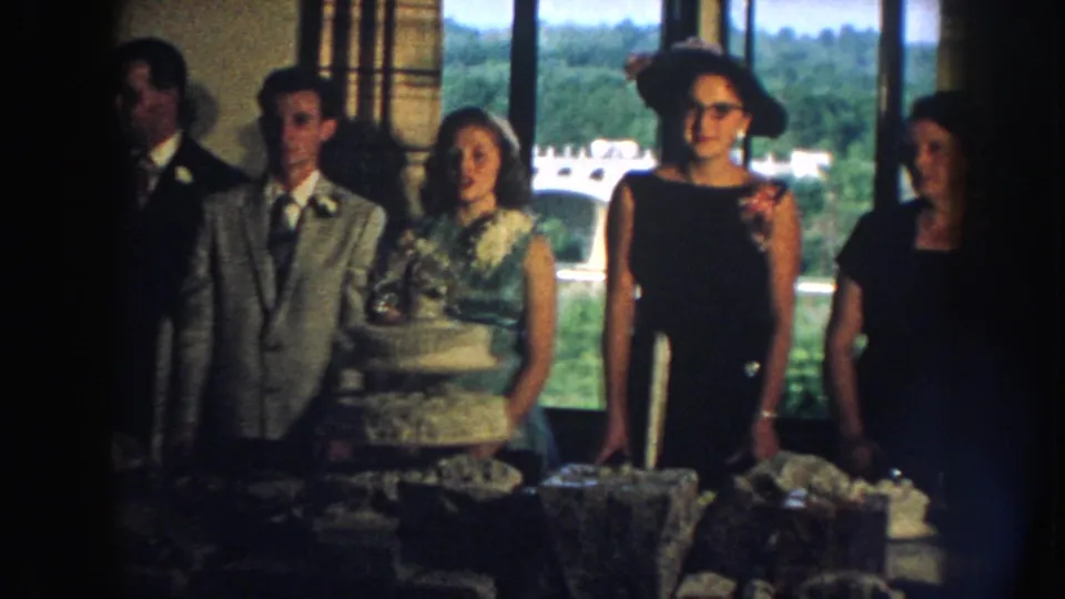 1959 Nebraska wedding reception frame with a posed group, tiered cake, formal clothing, and greenery visible beyond the windows.