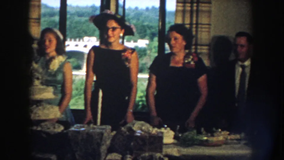 1959 Nebraska home-movie frame showing wedding guests posed beside the cake table in a private indoor reception setting.