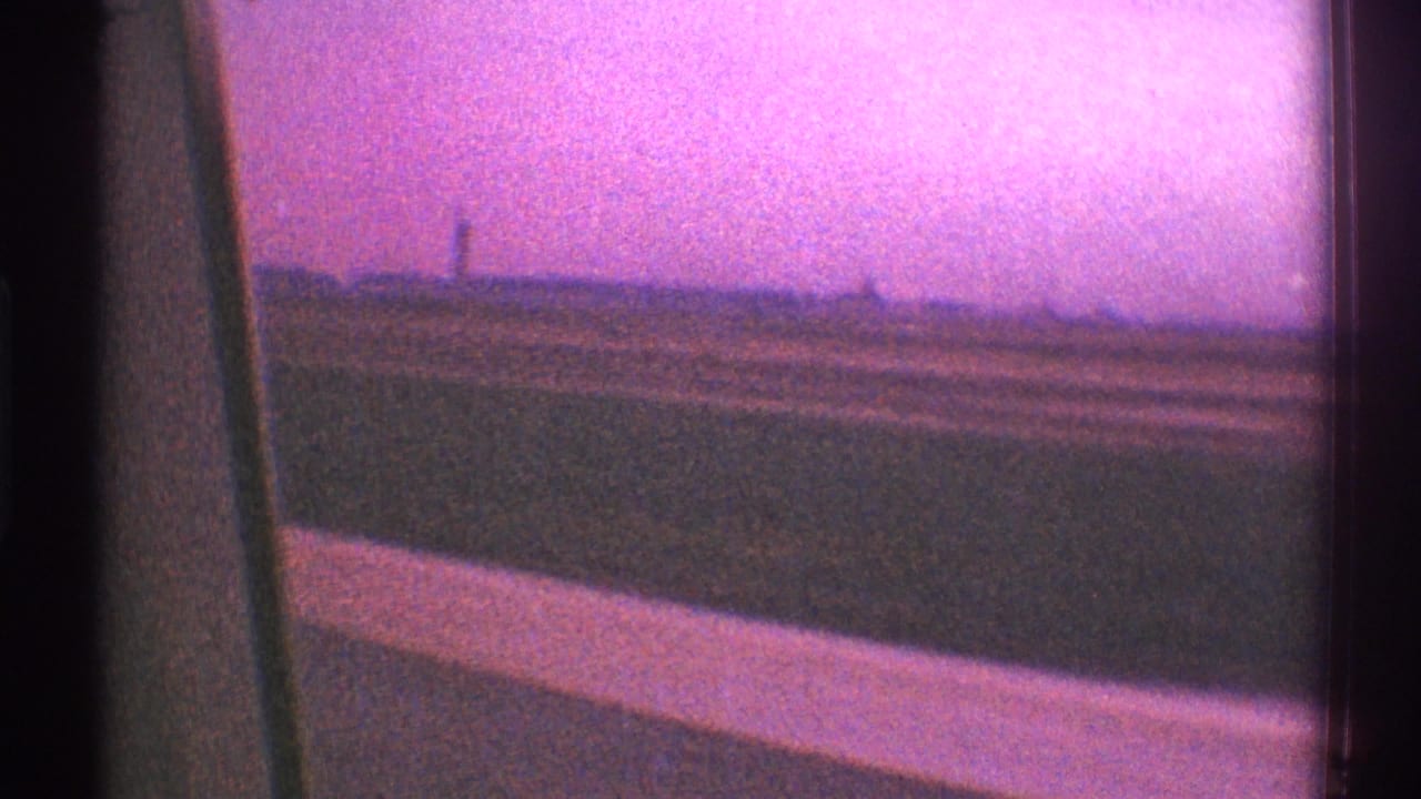 1969 Los Angeles home-movie frame looking from a moving vehicle past a dark window edge toward low grass, horizon, and purple-pink sky.