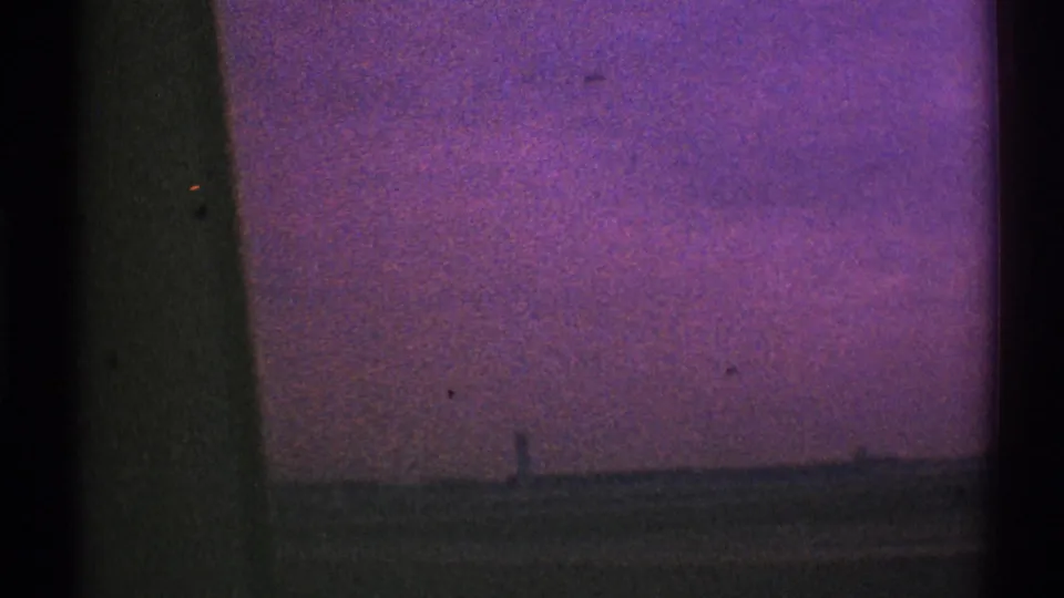 Final 8mm frame from the Los Angeles driving view, with a dark window pillar, low grassy horizon, and purple-pink sky.