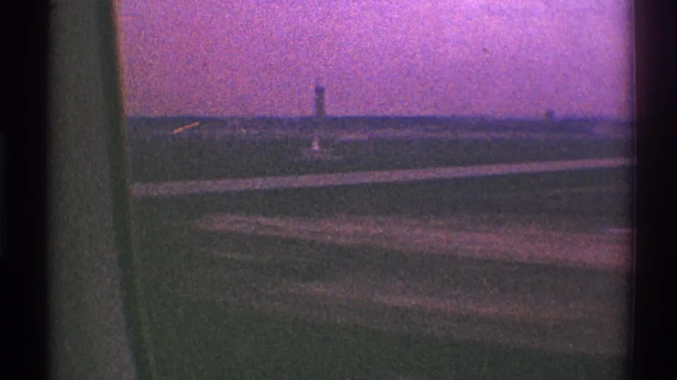 Passenger-window view over Los Angeles airport grounds in 1969, with runway pavement, grass, and a distant tower on the horizon.