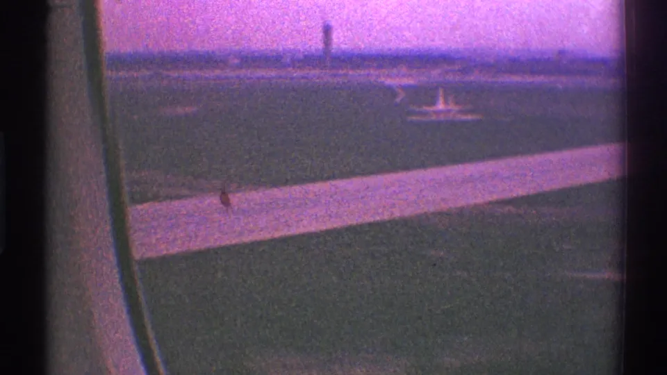 Aircraft wing and Los Angeles airport runway area seen from a passenger window, with a distant control tower under a color-shifted sky.