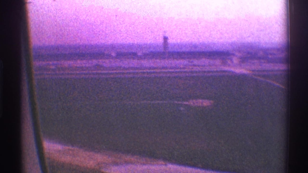 1969 Los Angeles airplane-window view over open fields, distant low development, and a tower-like structure in purple-tinted 8mm footage.